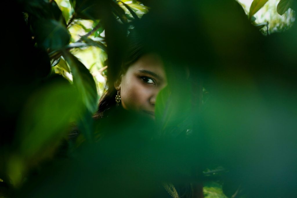 A woman partially hidden within lush green leaves, creating a sense of mystery.